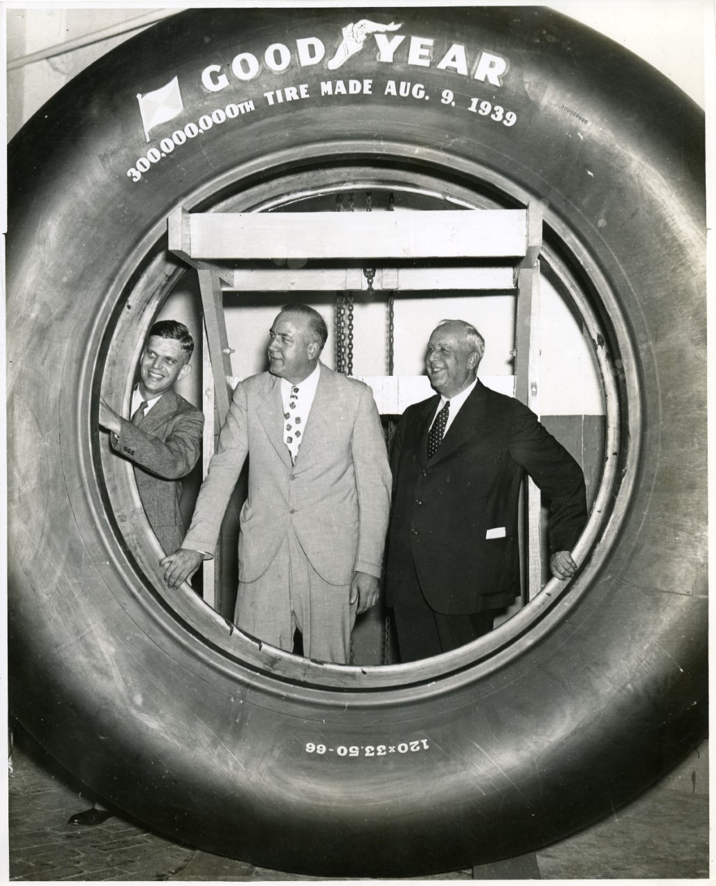 Three men in suits standing behind a very large Goodyear tire, which frames them. Text on the tire reads "300,000,000th tire made August 9, 1939."