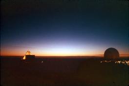Both top and bottom of the image are black. In the middle of the image is the last sliver of the sun on the horizon. The edges of the horizon are blue and orange. On the left there is a lit-up triangle structure and on the right is a circle structure