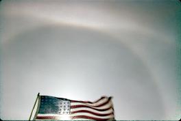 The arc appears at the zenith, which is the highest point in the sky. The photo is taken looking up at the center of the arc. An American flag is blowing in the wind at the bottom of the image.