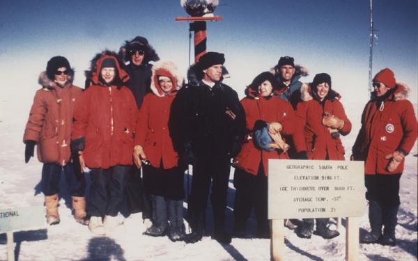 A group of women stand to take a photo at the south pole. The majority of the group is wearing heavy red coats. There is one individual in the center wearing a black coat. 