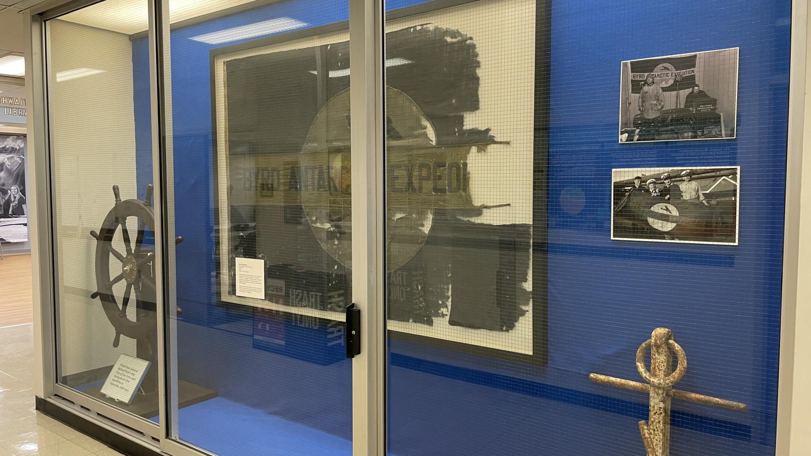 A side view from right of a full length built in display case outside of the Goldthwait Polar Library including a ship's large white anchor, a ship's steering wheel in dark wood and a large framed fabric shredded flag and 2 side images on a blue background behind a glass display