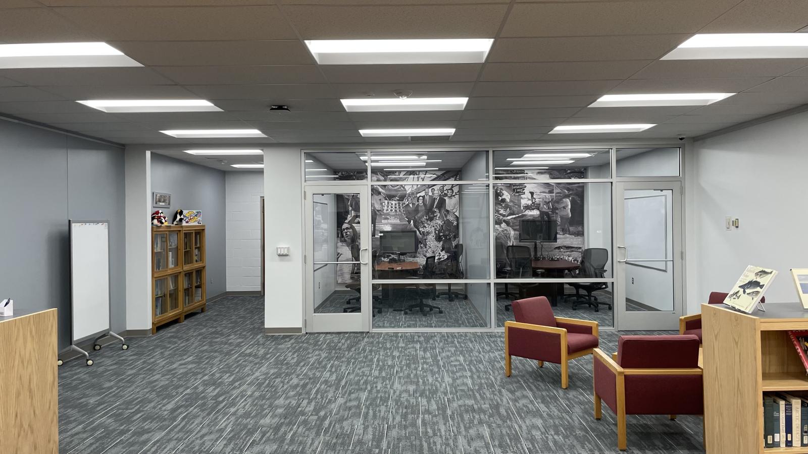 image taken from inside the Goldthwait Polar Library in the far back 2 glass enclosed quiet rooms  with a wall length vinyl drape of images on the back wall with some chairs and desk inside and gray carpet, white walls and a light wood table and 2 lounge chairs with red fabric in foreground on bottom right  and book shelve and white portable erase board on left side and white tile drop ceilings with with squares and come containing florescent light fixtures
