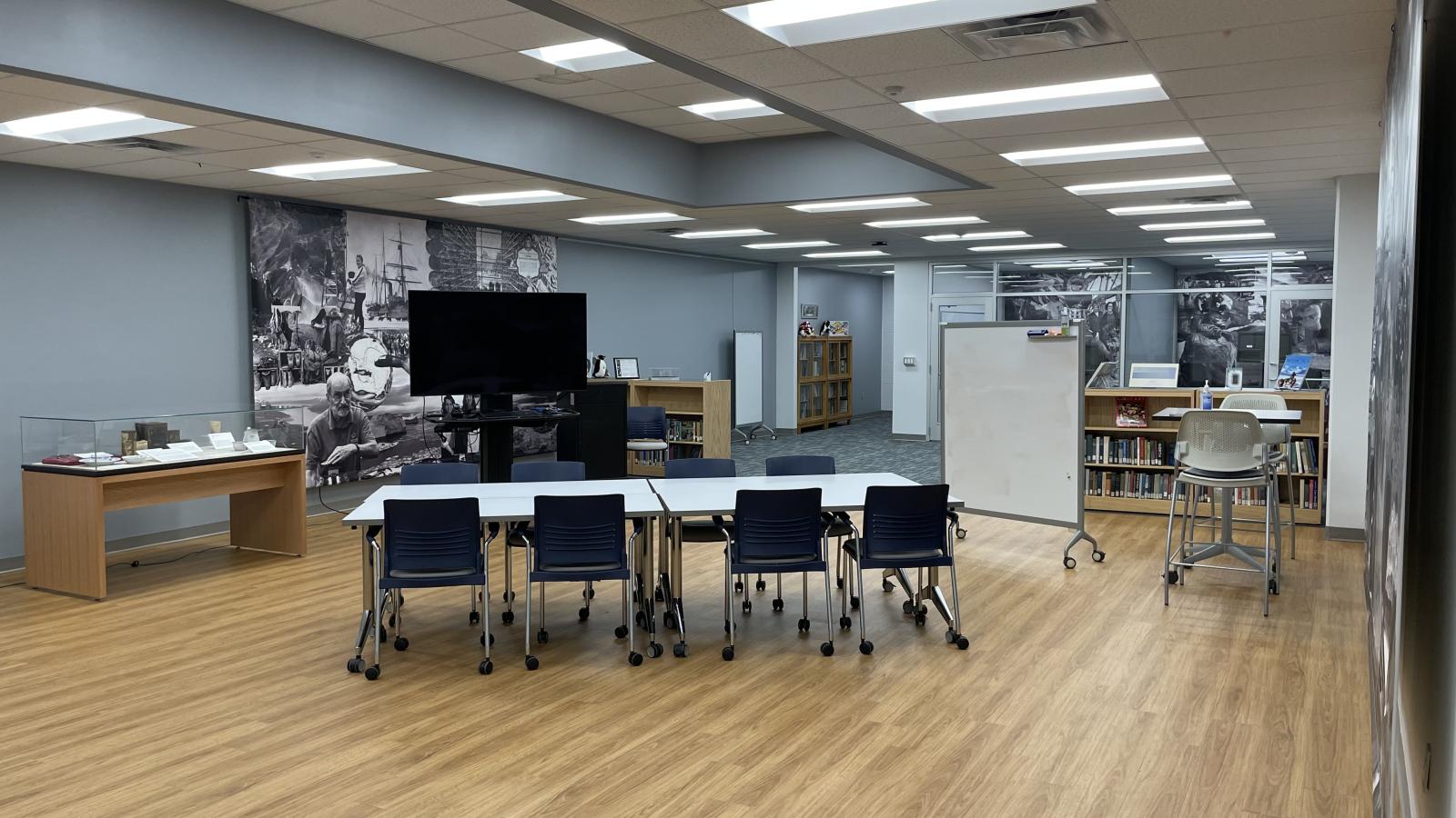 image taken from inside the Goldthwait Polar Library with white table and rolling chairs around it in the middle of the room with blue walls and a vinyl wall length drape image and a large monitor on rolling wheels and a display case next to it on light wooden base with light wood floors and light book shelve and white portable dry erase board in center behind table and white tile drop ceilings with with some squares containing florescent light fixtures that are on