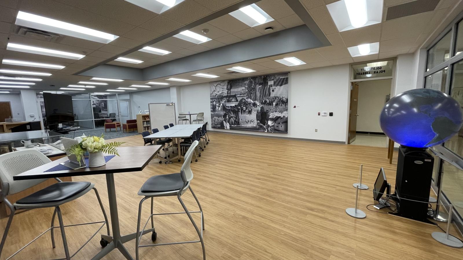 inside the Goldthwait Polar Library with white table and rolling chairs in the center with white walls and a vinyl wall length drape image on right with a large lit glob on a black stand next to glass wall and a large monitor next to a glass display case on light wooden base to the left with light wood floors and light book shelve and white portable dry erase board behind table and white tile drop ceilings with with some squares containing florescent light fixtures that are on