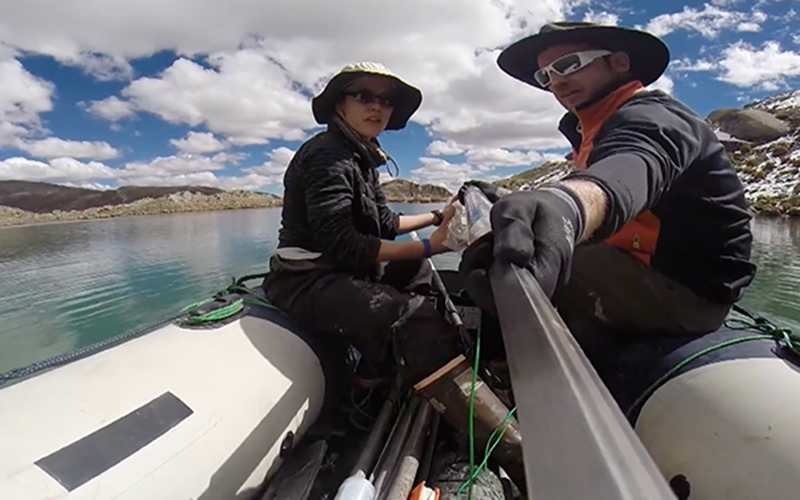 Two researchers are on a small raft on a large body of water. They are wearing hats, sunglasses, and water ready outdoorsy gear.