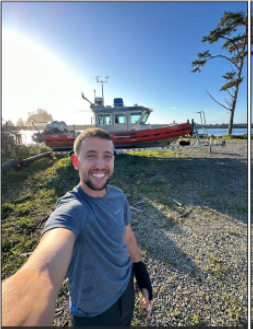 James Roubal taking a selfie outdoor with a backdrop of a boat, water and grass on a sunny day.