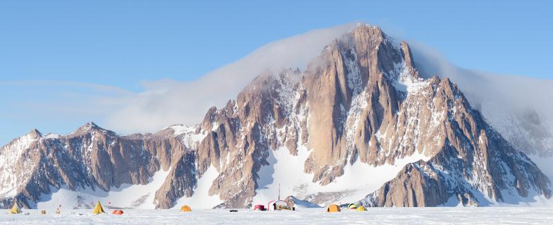 Mount Tidd in the Pirrit Hills in Antarctica with a number of different shape tents at its base under blue skies