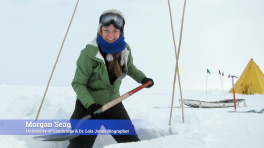 A white woman in snow gear with a shovel. She's standing in a shallow pit carved out of the surrounding snow. Behind her is a tent and a sled.