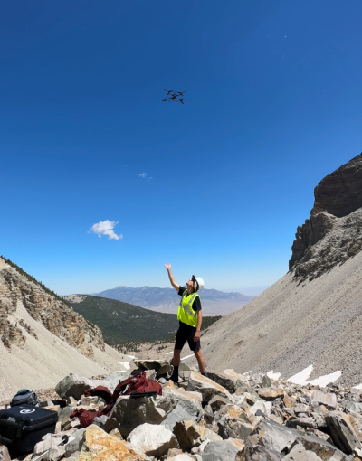 A person standing on rocks in a mountainous region with a yellow vest and hard hat looking up at a drone above his head with his hand reached out as if he just released the drone under blue skies
