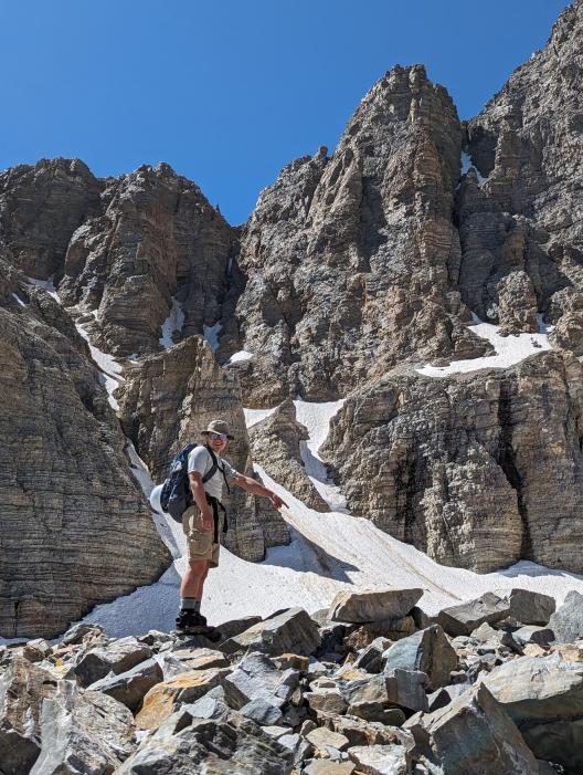 A person standing in mountains surrounded by rocks and snow pointing to a rock.