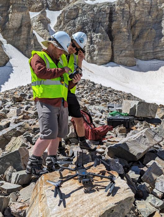 Two people in neon work vests and hard hats working on a drone in the mountainous field surrounded by rocks.