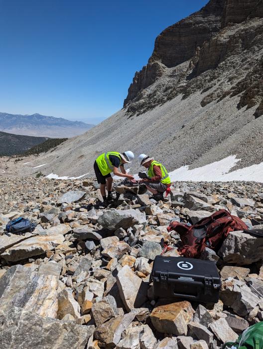Two people in hard hats and neon vests bent over working on a drone in the mountains with some snow covered ground.