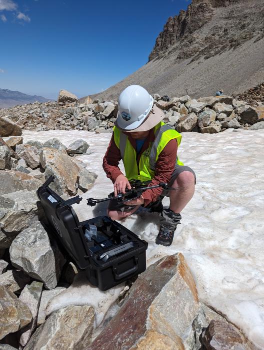 A person in a hard hat and neon work vest knelt down over a black toolbox working on a drone in the mountains with snow covered ground.