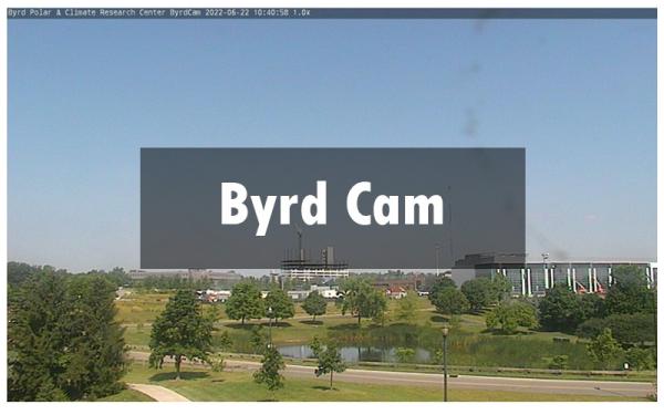 Still image from the Byrd Center weather camera taken during summer. The camera overlooks a park with a pond. There are buildings in the distance. Overtop is the text "Byrd Cam"
