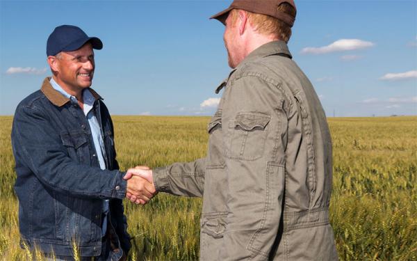 Two white men shaking hands in a wheat field. Both are wearing baseball caps and jackets. The one to the left is wearing blue jean and smiling. The one on the right is wearing brown canvas. His face is not visible.