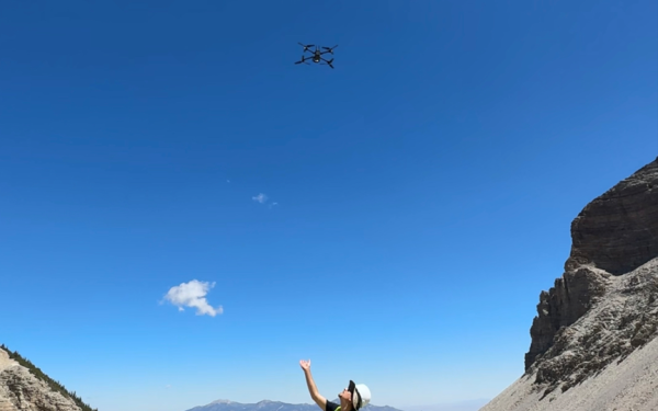 A person standing on rocks in a mountainous region with a yellow vest and hard hat looking up at a drone above his head with his hand reached out as if he just released the drone under blue skies