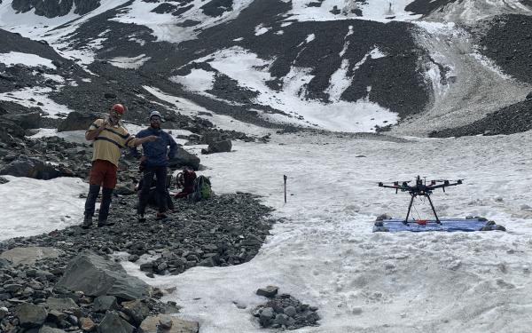 Two people posing for the camera on a rocky, snow covered mountain with a drone. 