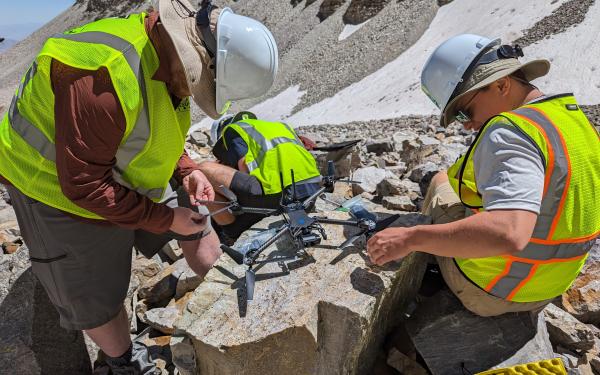 Three people in neon work vests and hard hats working on a drone in the mountainous field surrounded by rocks.